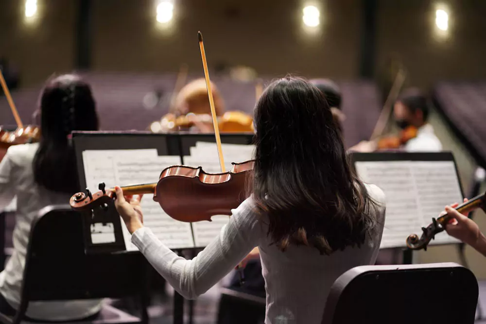 Photo of students playing violins and reading sheet music. Their backs are to the camera and the. audience is visible. Image by Milk Tea on Unsplash.